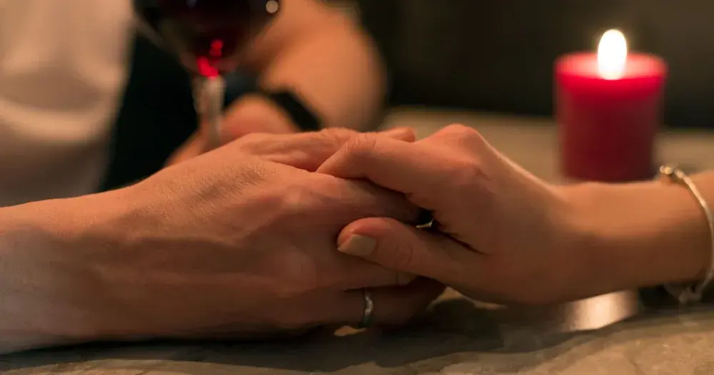 Mãos de um casal entrelaçadas sobre a mesa, à luz de vela, com uma taça de vinho ao fundo