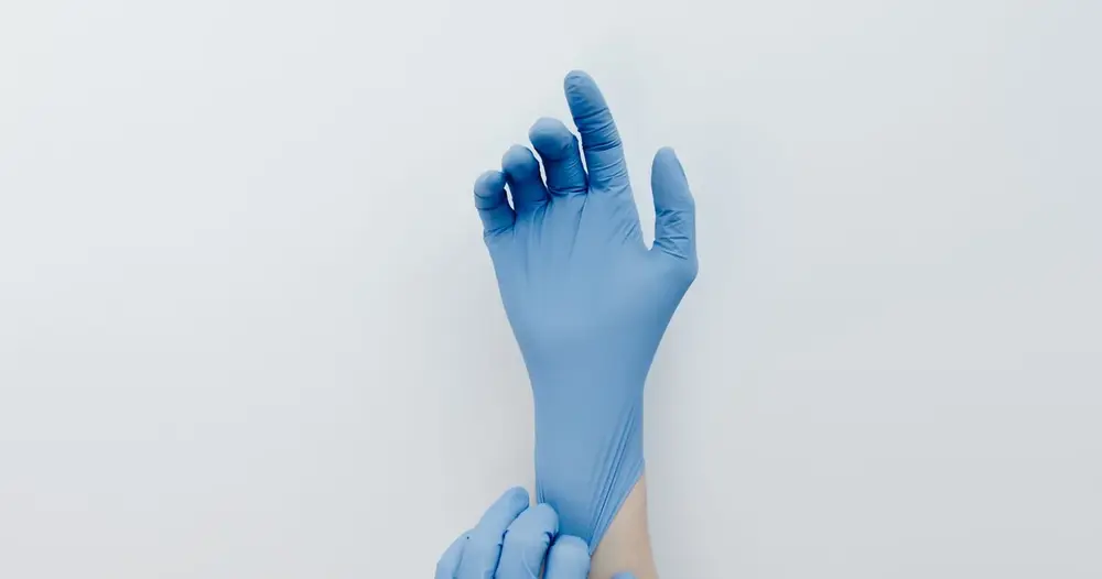 Mão calçando luva azul de procedimento sobre fundo claro, exemplo de luvas de procedimento