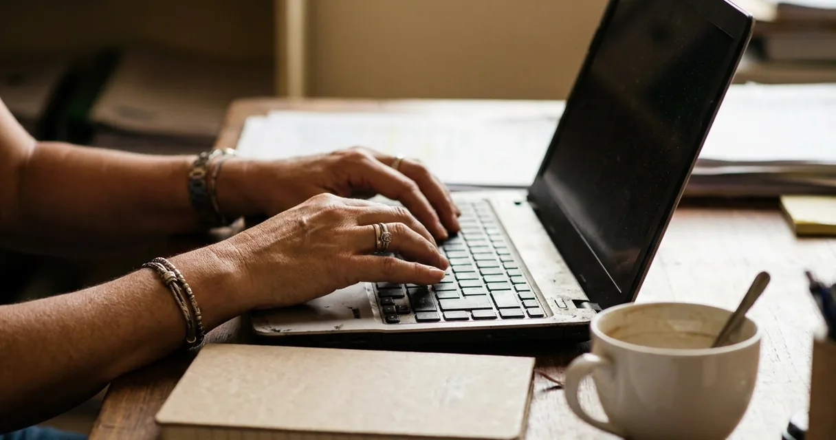 Mãos digitando em um notebook sobre mesa de madeira, com caderno fechado e xícara de café ao lado